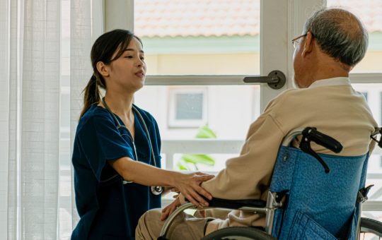 Caring nurse with senior man sitting on wheelchair at home.