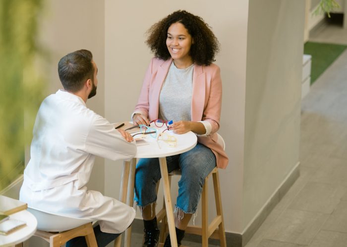 Pretty young client of optics shop or clinics sitting by table in front of clinician consulting her about choice of eyeglasses