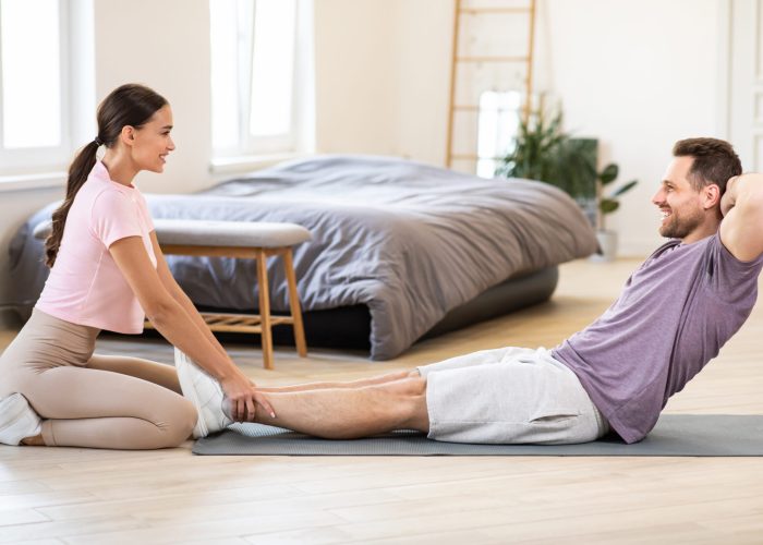 Sporty Couple Exercising Together Training Doing Abs Sit-Ups At Home, Girlfriend Holding Boyfriend's Legs Helping Him Exercise. Fitness Routine For Two Concept